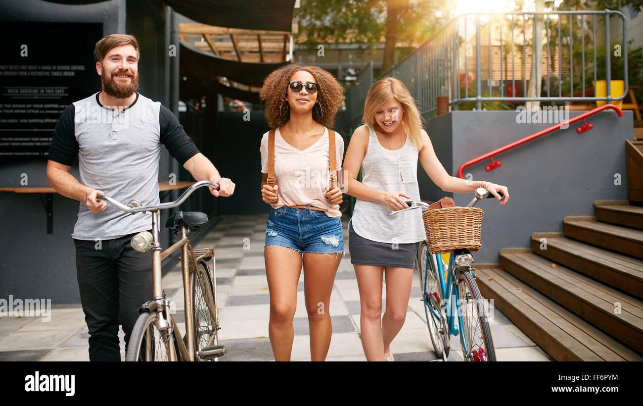Trois jeunes gens marchant dans la rue avec leur vélo. Les amis masculins et féminins sur route avec leur vélo. Banque D'Images Trois jeunes gens marchant dans la rue avec leur vélo. Les amis masculins et féminins sur route avec leur vélo. Banque D'Images