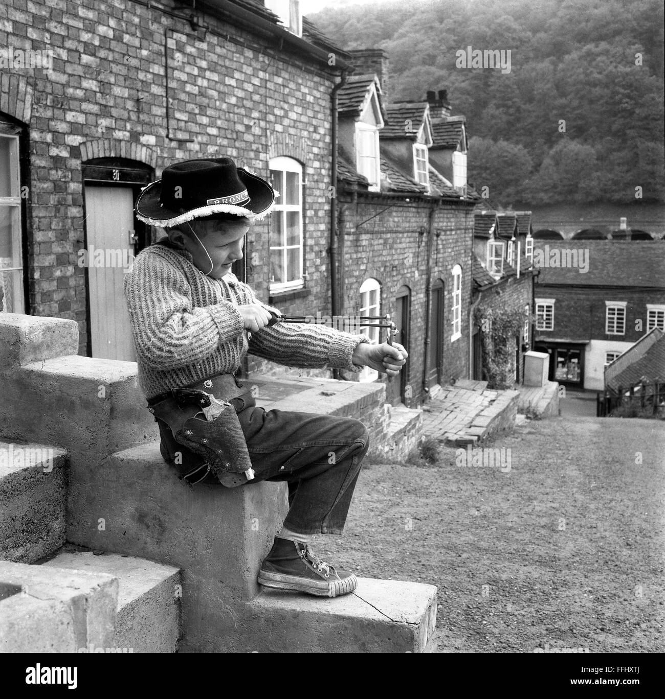 Jeune garçon jouant seul avec une catapulte et portant une tenue de cowboy 1960s Grande-Bretagne rue à l'extérieur des rues Banque D'Images