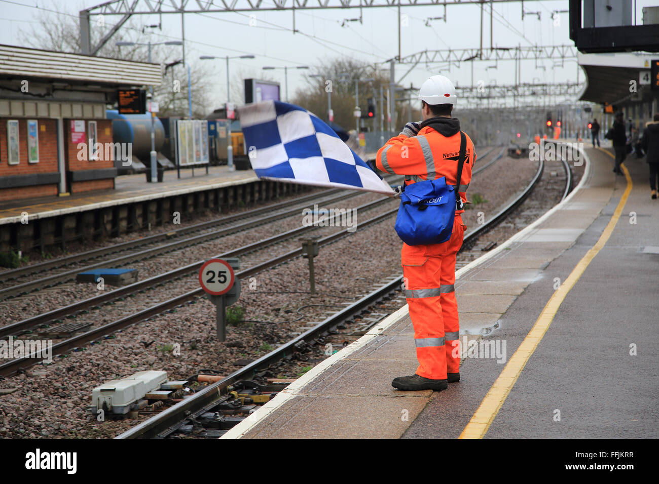Employé des chemins de fer portant des vêtements haute visibilité orange tient à carreaux bleu et blanc du drapeau de sécurité, Colchester, Essex, Angleterre, Royaume-Uni Banque D'Images