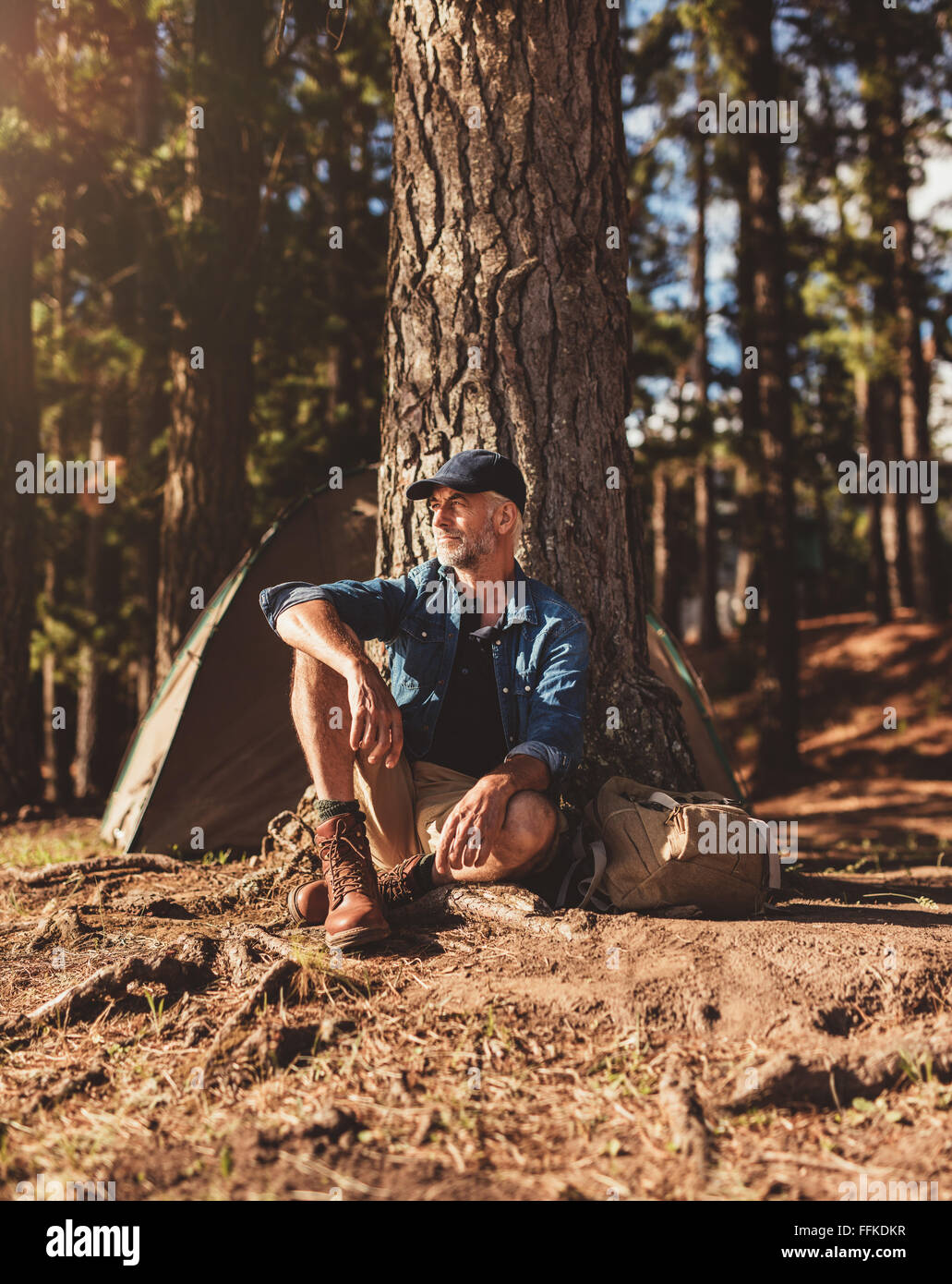 Portrait of senior man assis tout seul par un arbre avec une tente dans l'arrière-plan. Homme mûr assis dans un camping à l'écart sur summ Banque D'Images