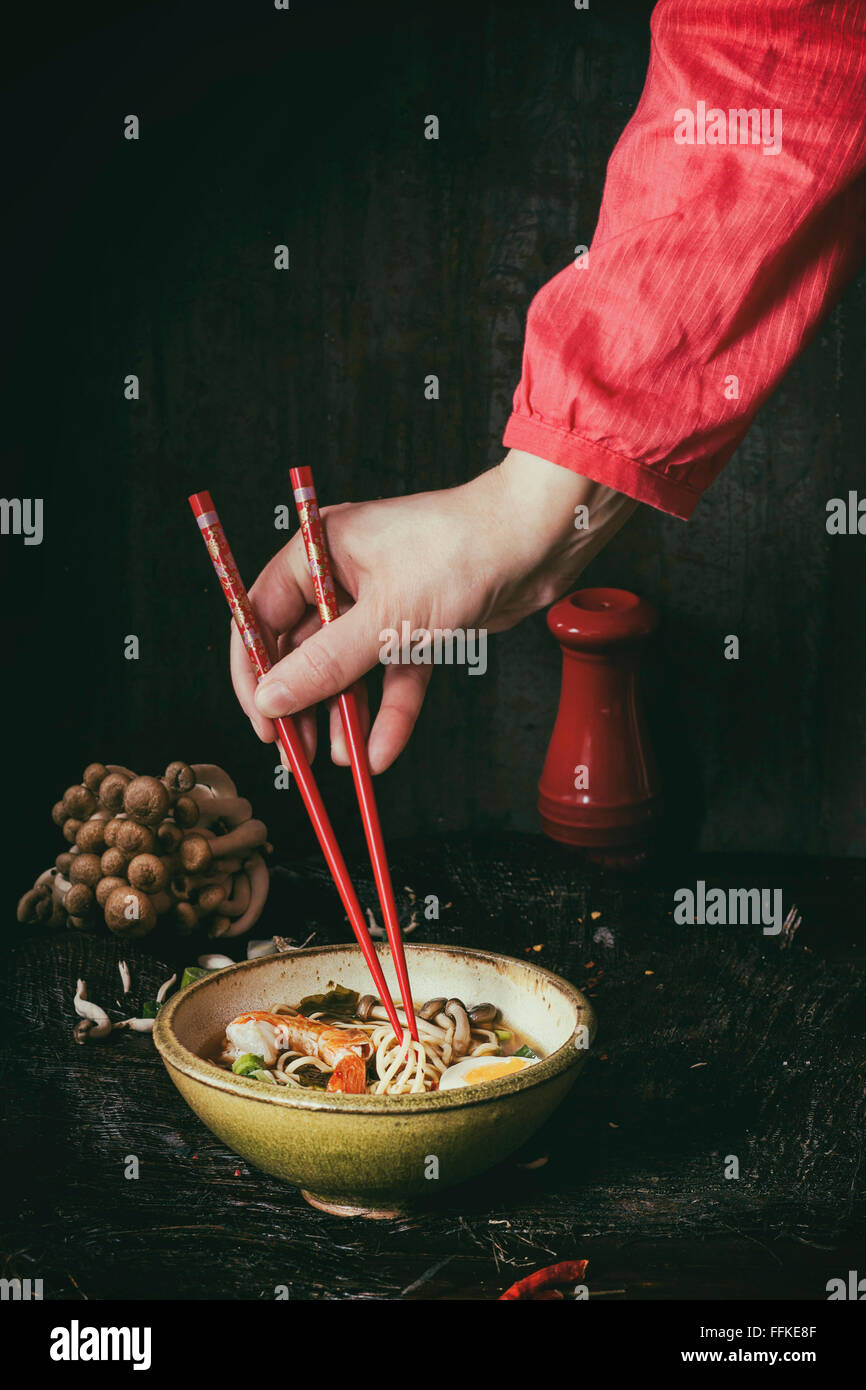 En main de femme chemise rouge prendre par baguettes rouge de nouilles ramen Soupe Asiatique aux crevettes, l'oignon, des tranches de champignons et d'œufs, la serv Banque D'Images