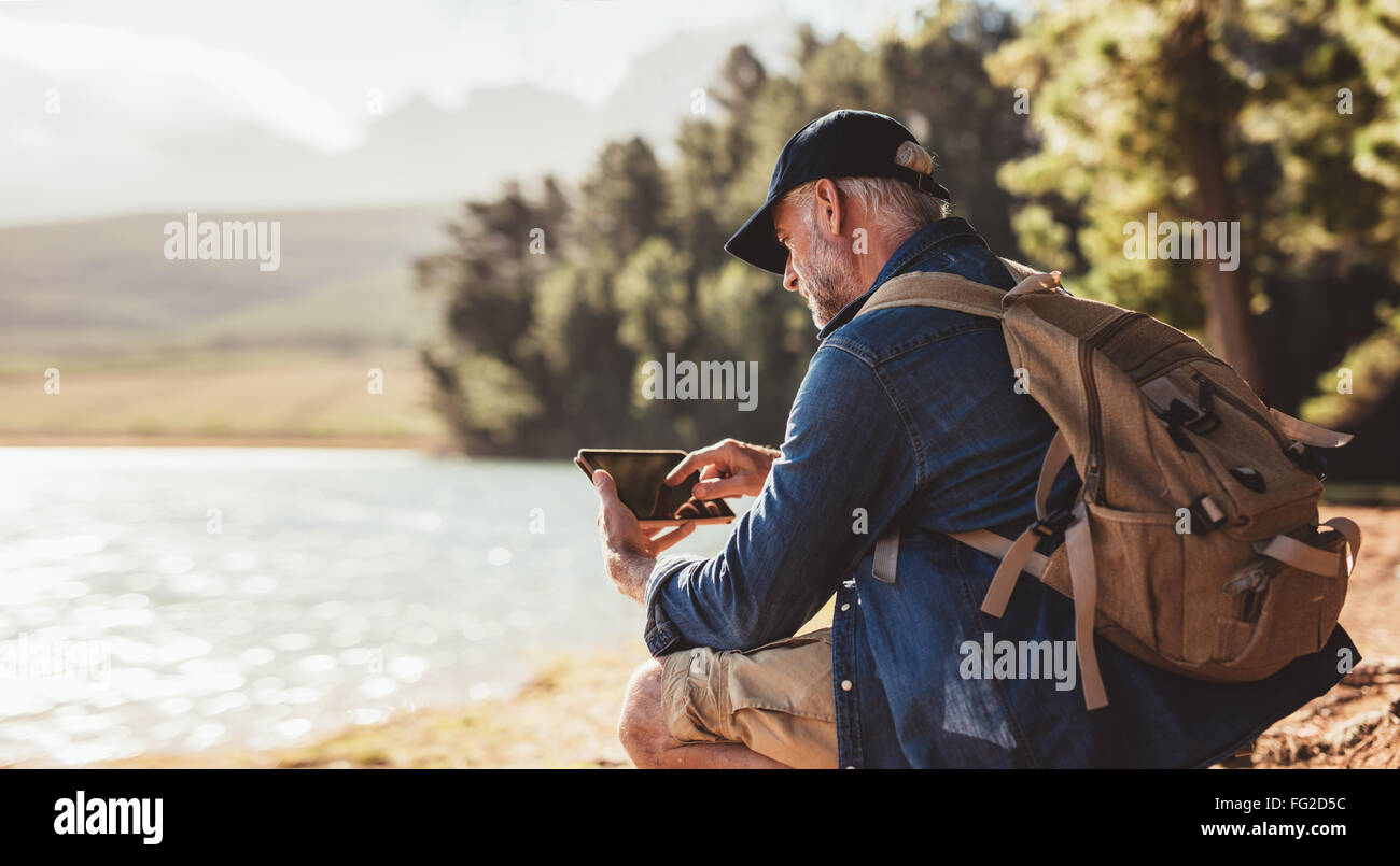 Man avec sac à dos en utilisant l'onglet numérique tout en étant assis près d'un lac. Homme mûr sur la randonnée dans la nature à l'aide de tablette numérique. Banque D'Images