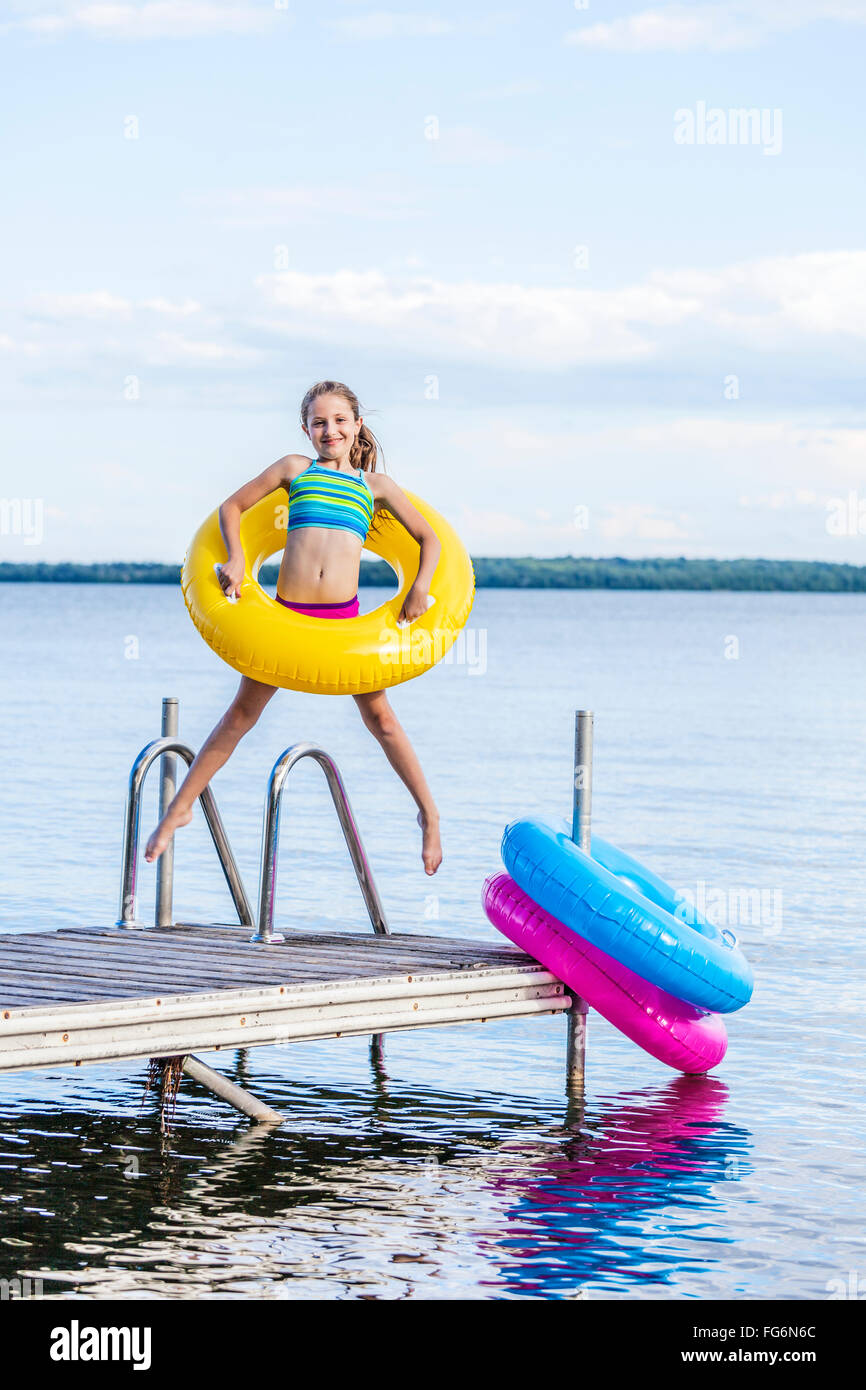 Girl jumping on dock en Lac Balsam tenant un anneau gonflable jaune ; Ontario, Canada Banque D'Images