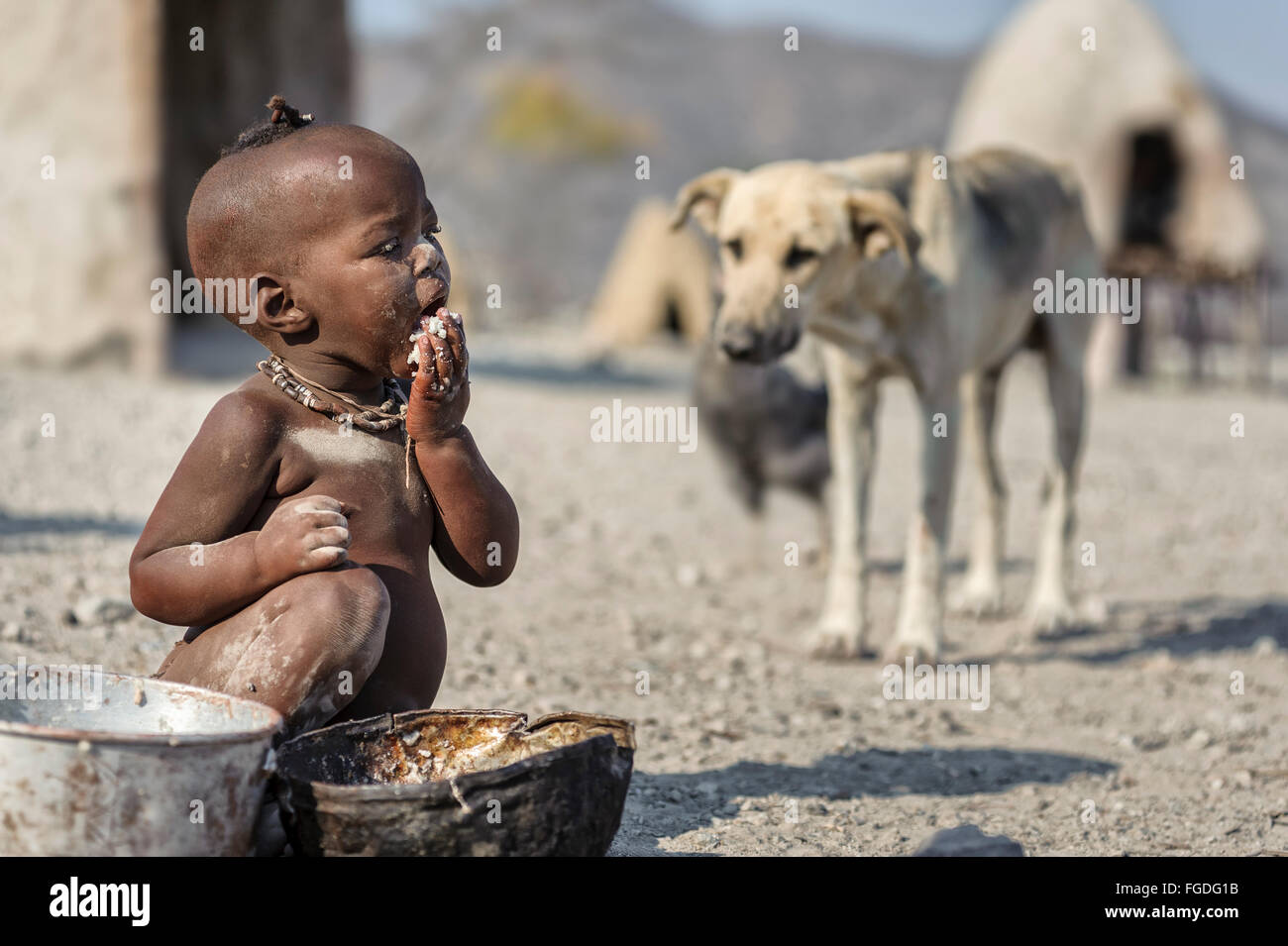 Enfant Himba assis sur le sol de son village éloigné de manger un pot rouillé tandis qu'un chien est à proximité de lui. Banque D'Images