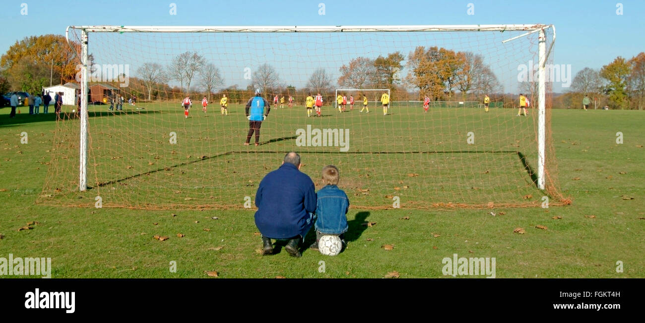 Père & Fils à balle derrière poteau de but regardant teen kids playing football match village jeu de soccer père vivre ensemble avec collage enfant UK Banque D'Images