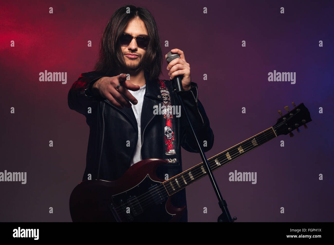 Jeune homme séduisant chanteur avec guitare holding microphone et pointant sur vous sur fond de fumée colorée Banque D'Images