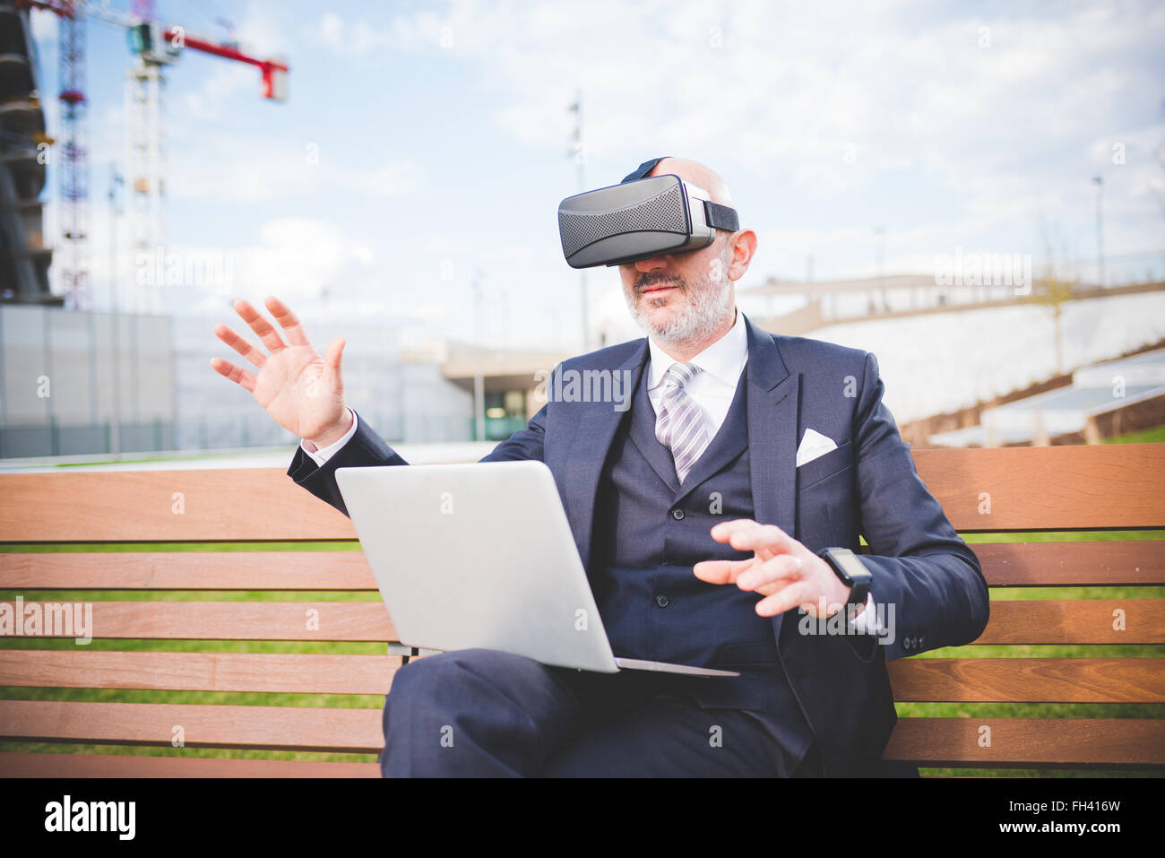L'âge moyen caucasian business homme assis sur un banc avec un ordinateur portable sur son genou en utilisant smartwatch et 3D viewer - futuristes, le multitâche, technologie concept Banque D'Images