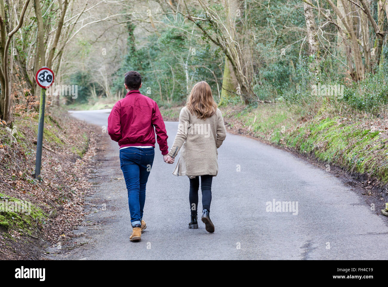 Jeune couple marche main dans la main le long d'une route de campagne bordée d'arbres au début du printemps, Surrey, Angleterre. Banque D'Images