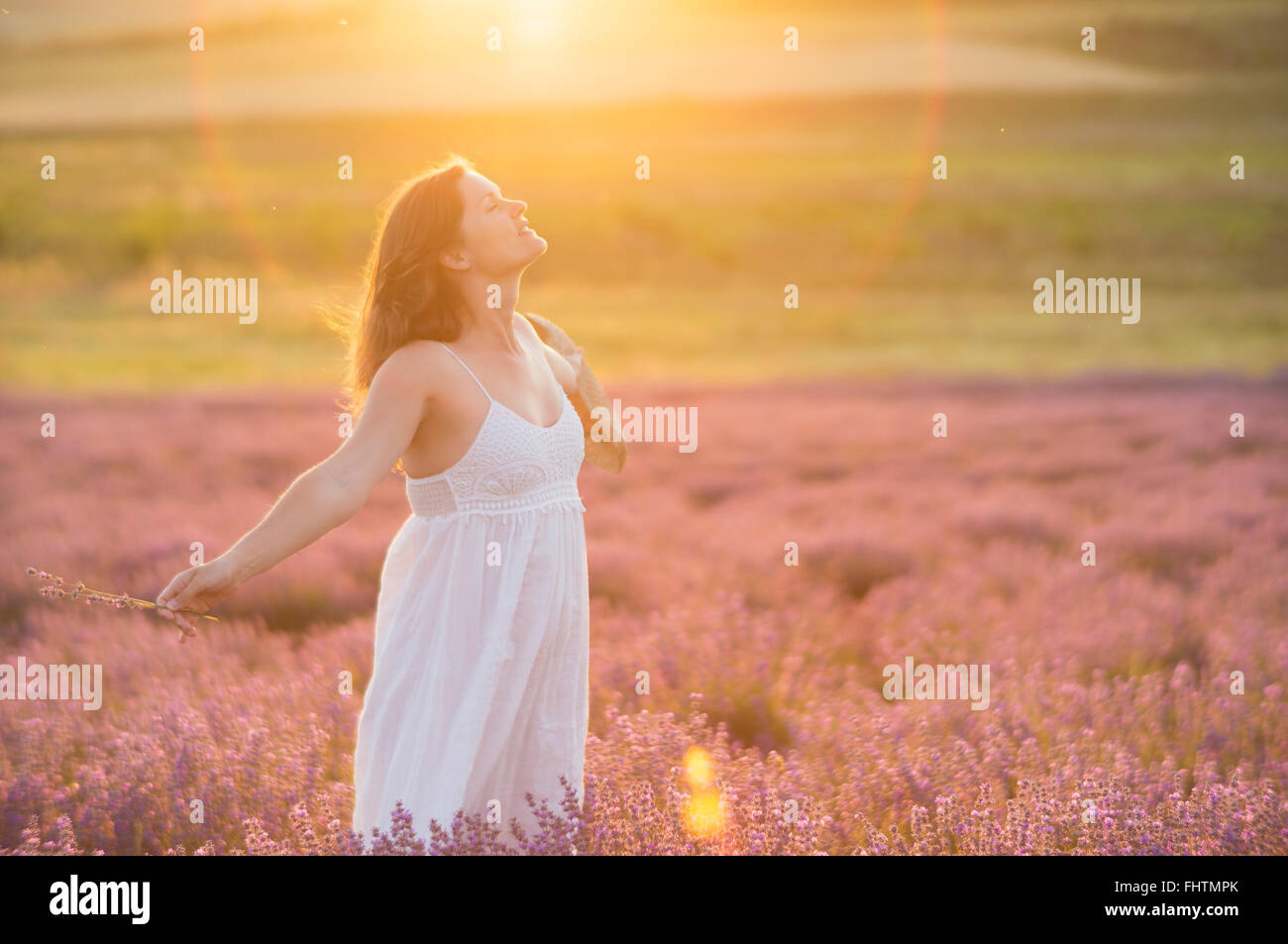 Belle jeune femme avec une robe blanche et un chapeau de paille, debout au milieu d'un champ de lavande à dans la lumière dorée de th Banque D'Images