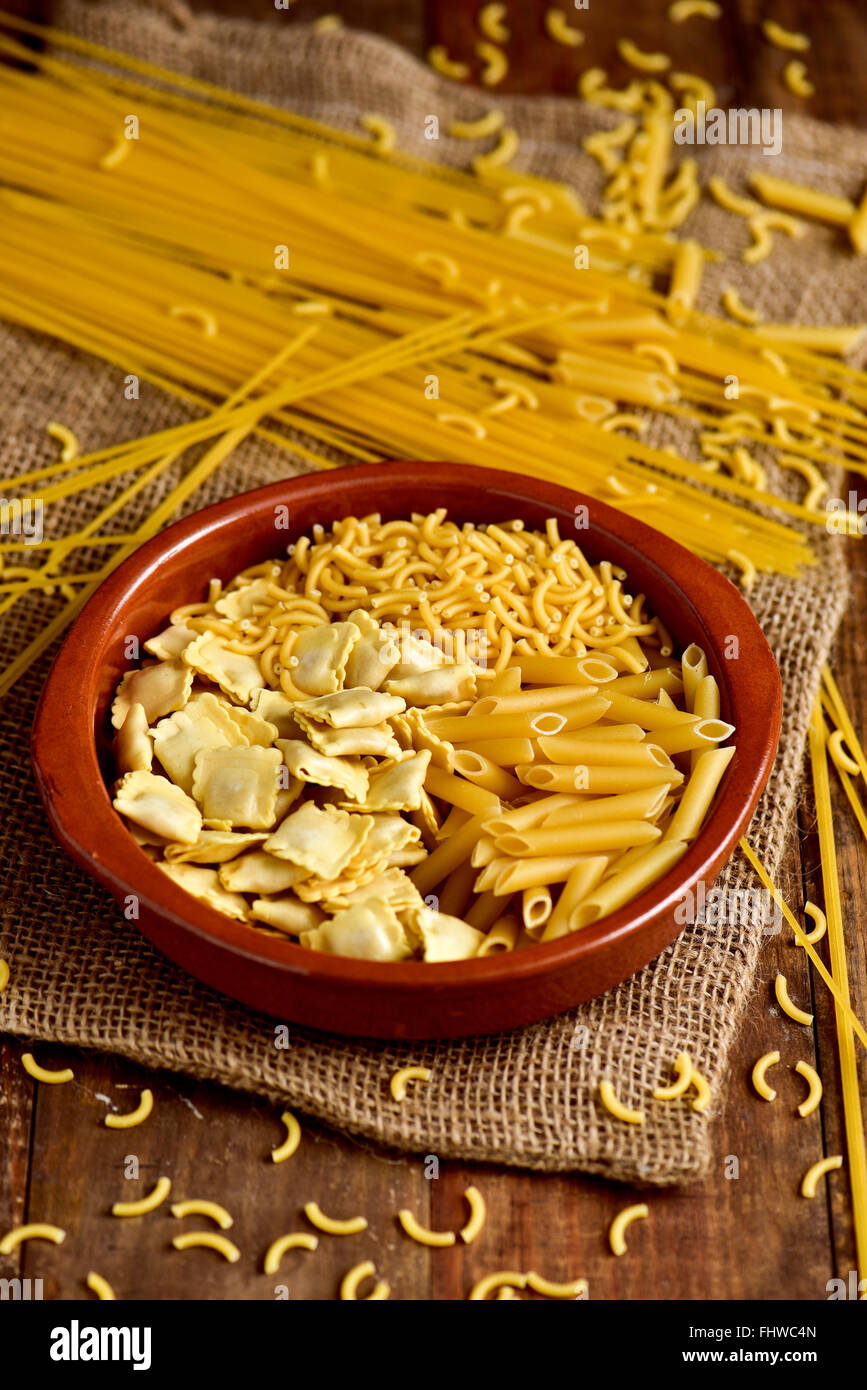 Libre d'un bol en terre cuite avec différentes pâtes alimentaires non cuites comme les ravioli, spaghetti ou mostaccioli, sur une table en bois rustique Banque D'Images