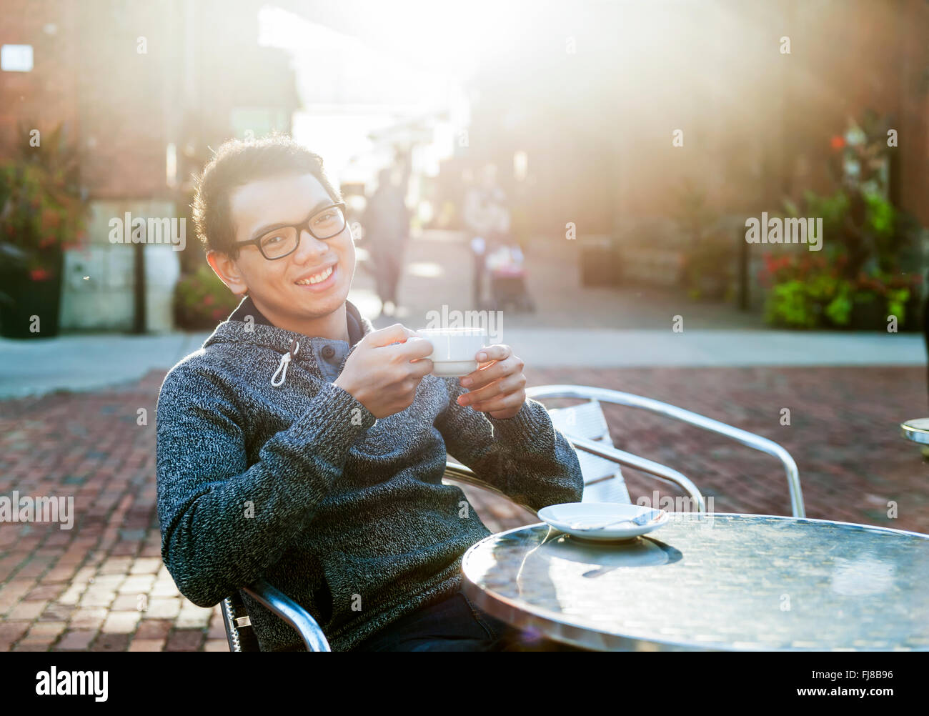 Young Asian man sitting and smiling in outdoor cafe holding tasse de café, des reflets. Banque D'Images