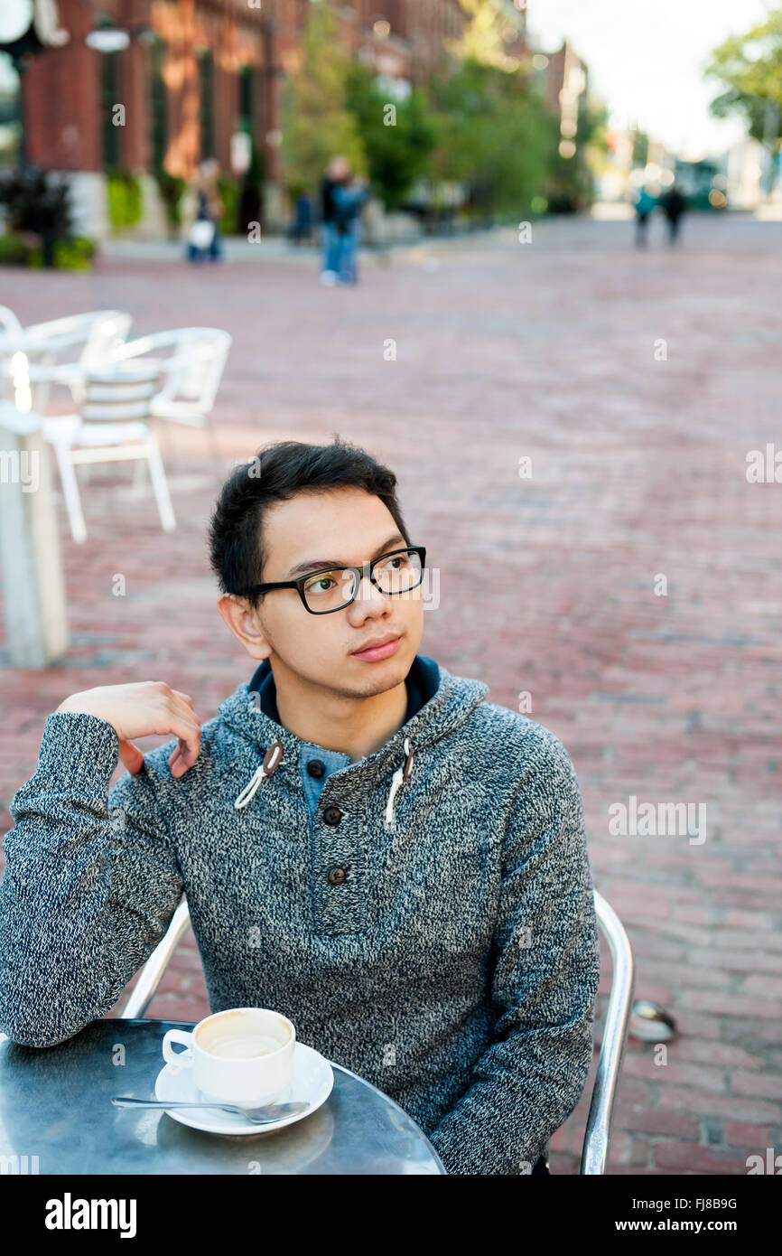 Young Asian man in outdoor cafe avec tasse de café à la suite de graves et contemplative Banque D'Images