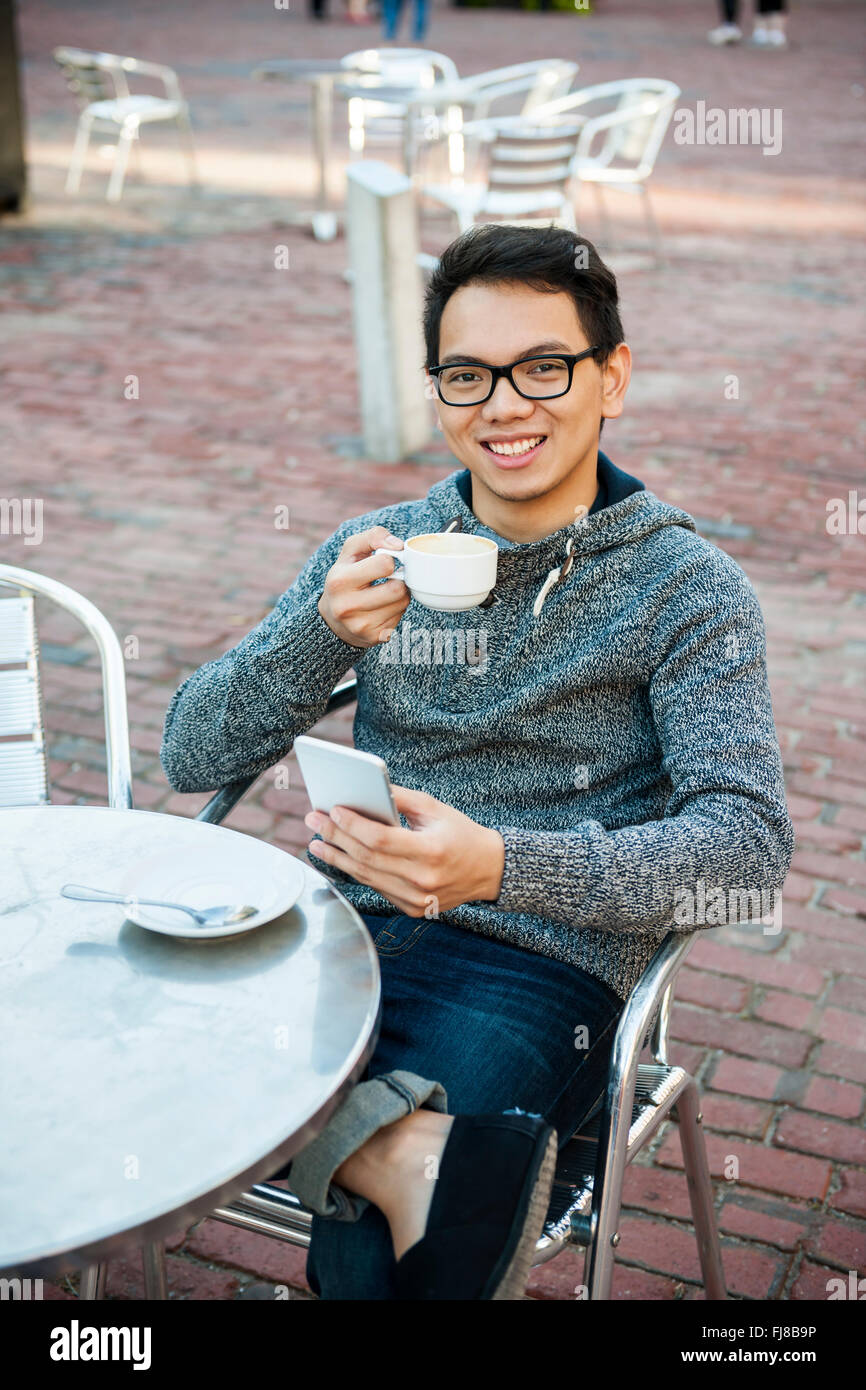 Young Asian man sitting and smiling in outdoor cafe holding relaxant tasse de café et la messagerie instantanée sur téléphone mobile Banque D'Images