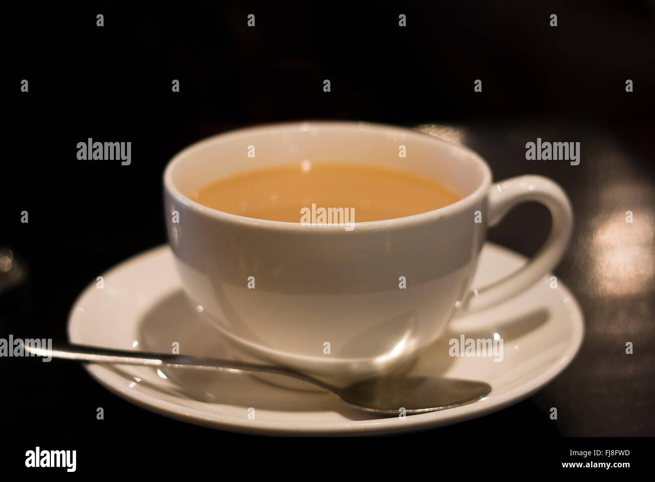 Une tasse de porcelaine remplie de café et crème assis sur une assiette avec une cuillère de métal au sommet d'une table dans un restaurant sombre Banque D'Images