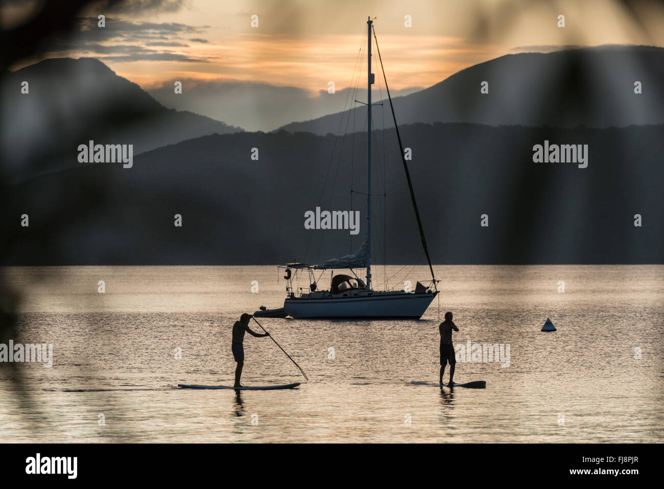 Silhouette de 2 stand up paddle boarders au coucher du soleil le long des côtes de l'île un paradis tropical en eaux calmes avec voiliers dans l'arrière-plan. Banque D'Images