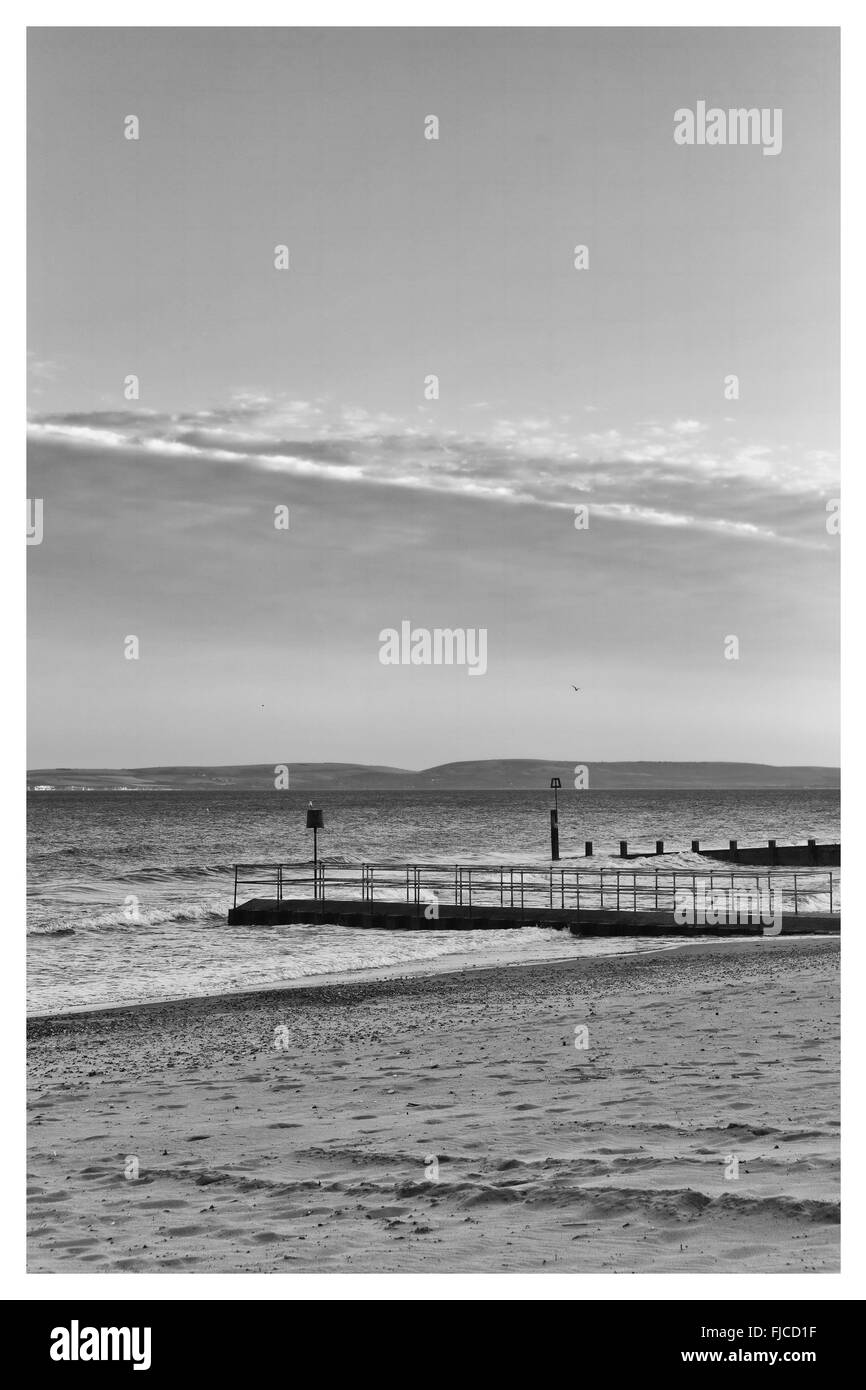 Une image en noir et blanc de la lumière au coucher du soleil d'un pilier à la direction de la mer à la plage de sable et une bande de clou spectaculaire Banque D'Images