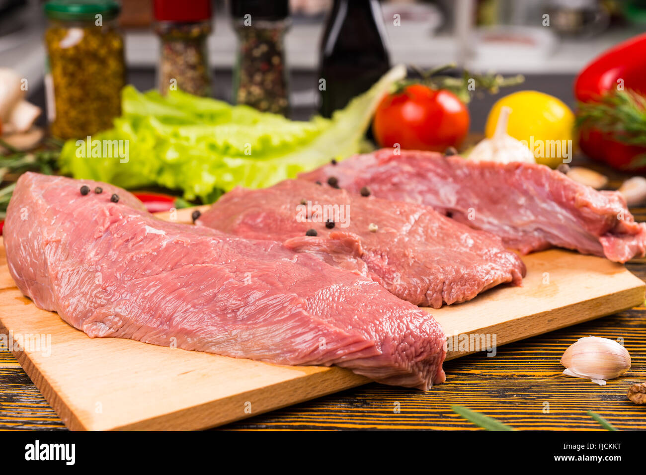 Deux grands morceaux de viande crue avec des poivrons sur une planche à découper en préparation pour la cuisson avec divers ingrédients de cuisine dans l'arrière-plan. Banque D'Images