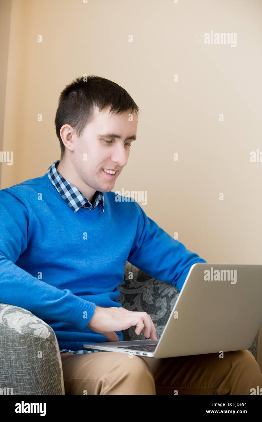 Portrait of smiling casual séduisant jeune homme assis dans un fauteuil à l'intérieur, à l'aide d'un ordinateur portable, regarder l'écran, homme Banque D'Images