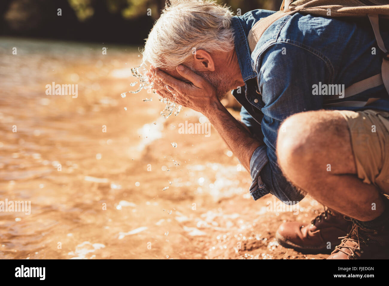Portrait de côté un homme mûr lave son visage dans le lac pendant une randonnée. Senior man with backpack assis au bord de l'eau un Banque D'Images