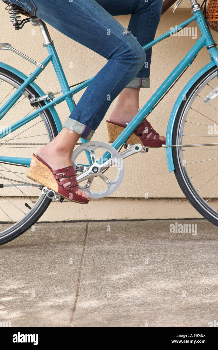 Les jambes de la femme un pédalage vélo sur un trottoir. Elle porte des jeans et des sandales à talons de coin. De vie sain mode été amusant dans la ville. Banque D'Images