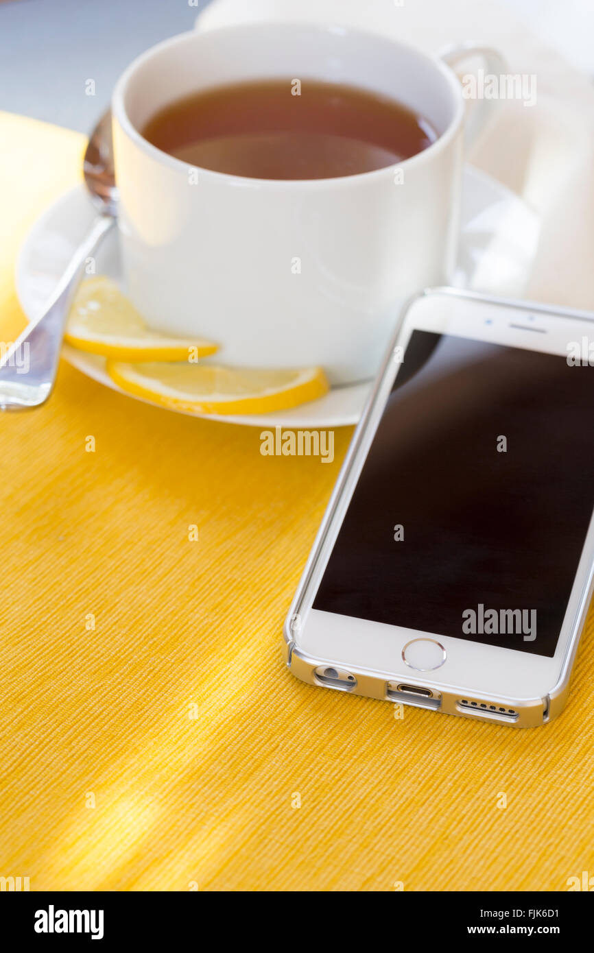 Tasse de thé au citron et de téléphones cellulaires avec écran noir assis sur table avec linge de table jaune. À l'aide de téléphone mobile au petit déjeuner le matin. Banque D'Images
