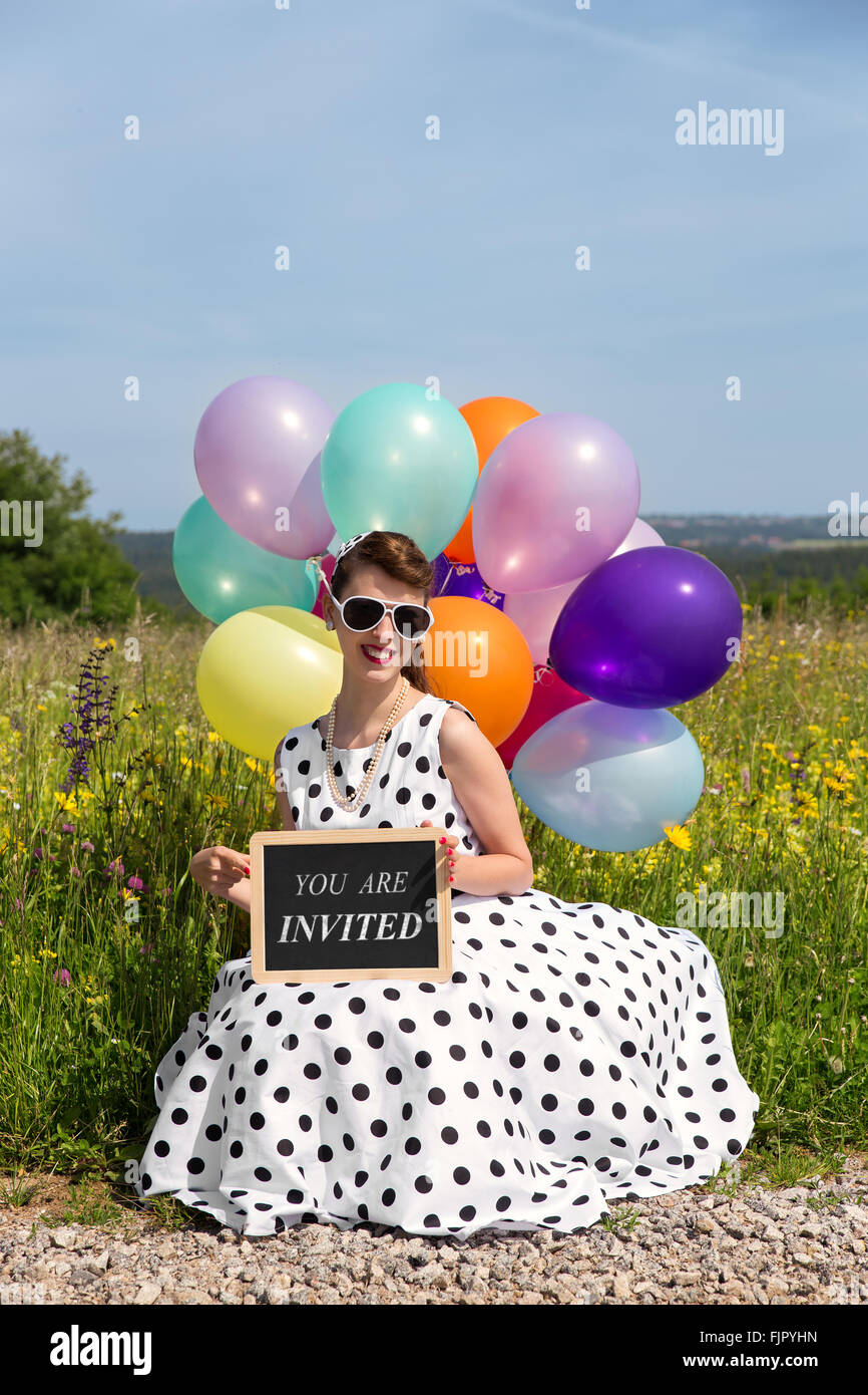 Les jeunes femmes dans une robe 50's ballons colorés et un conseil, la route par le texte que vous êtes invités, invitation concept Banque D'Images