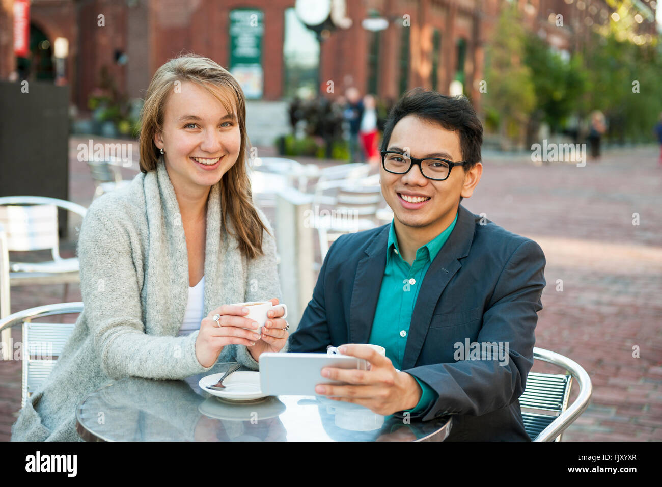 Deux jeunes gens souriants avec mobile device while sitting at outdoor cafe table on city street Banque D'Images