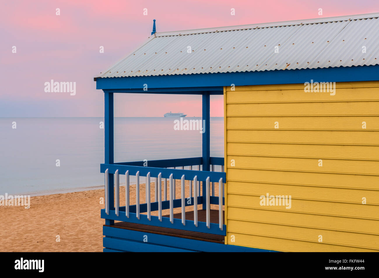 Cabane de plage jaune donnant sur un bateau de croisière à voile à travers l'eau au lever du soleil rose Banque D'Images