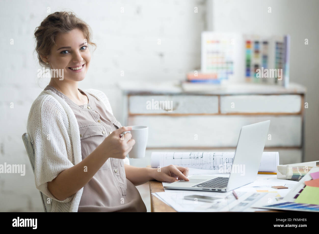 Portrait of beautiful happy smiling young designer woman sitting at home office 24 avec tasse de café, posing Banque D'Images