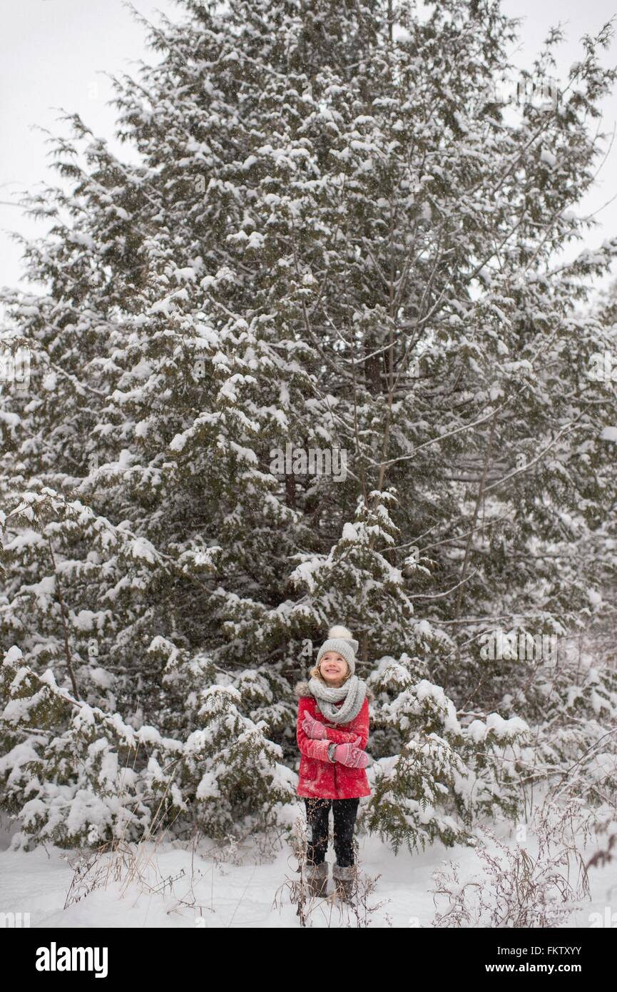 Jeune fille debout devant un grand arbre à feuilles persistantes, couvertes de neige Banque D'Images