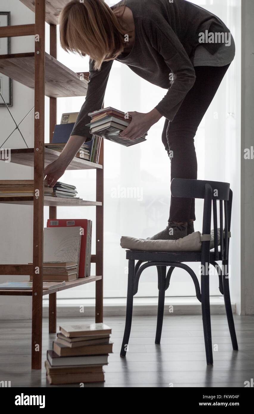 Femme debout sur une chaise pour organiser livres sur l'étagère de salon Banque D'Images