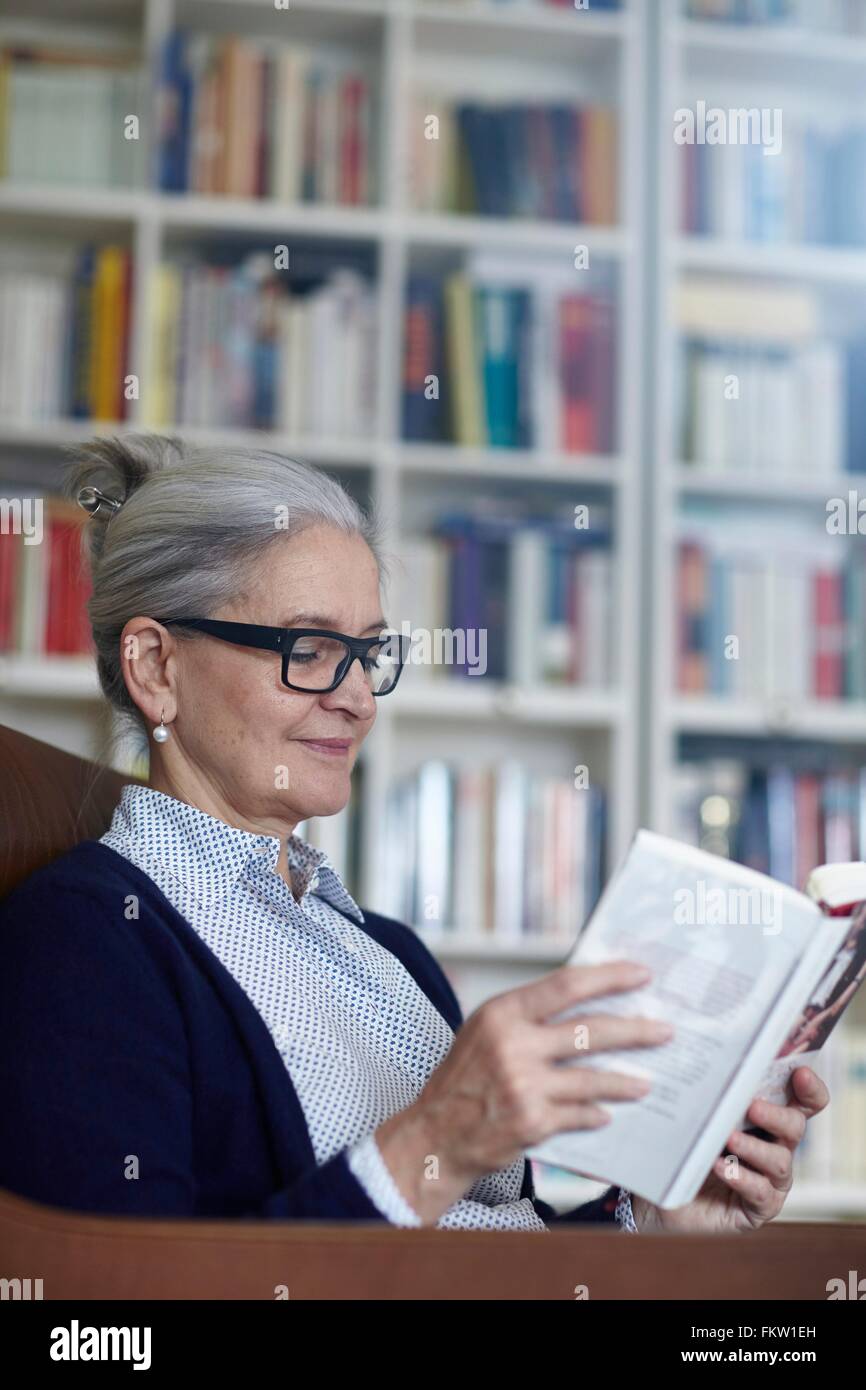 Grey haired young woman reading book from étagères Banque D'Images