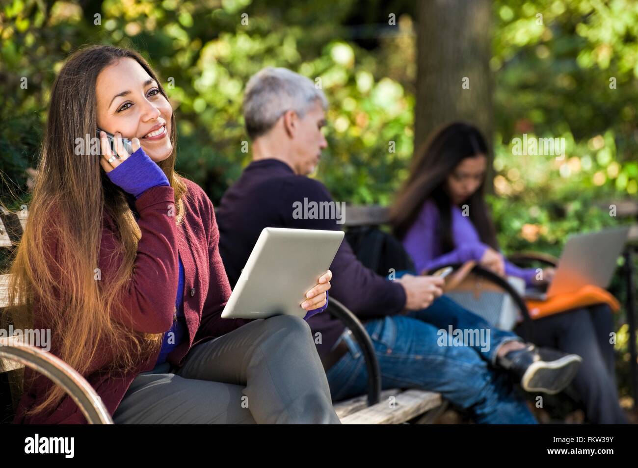 Young woman sitting on park bench using digital tablet et bavardant sur smartphone Banque D'Images