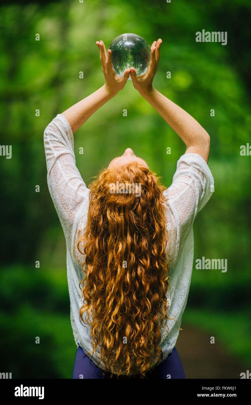 Vue arrière du jeune femme aux longs cheveux rouges holding up crystal ball in forest Banque D'Images