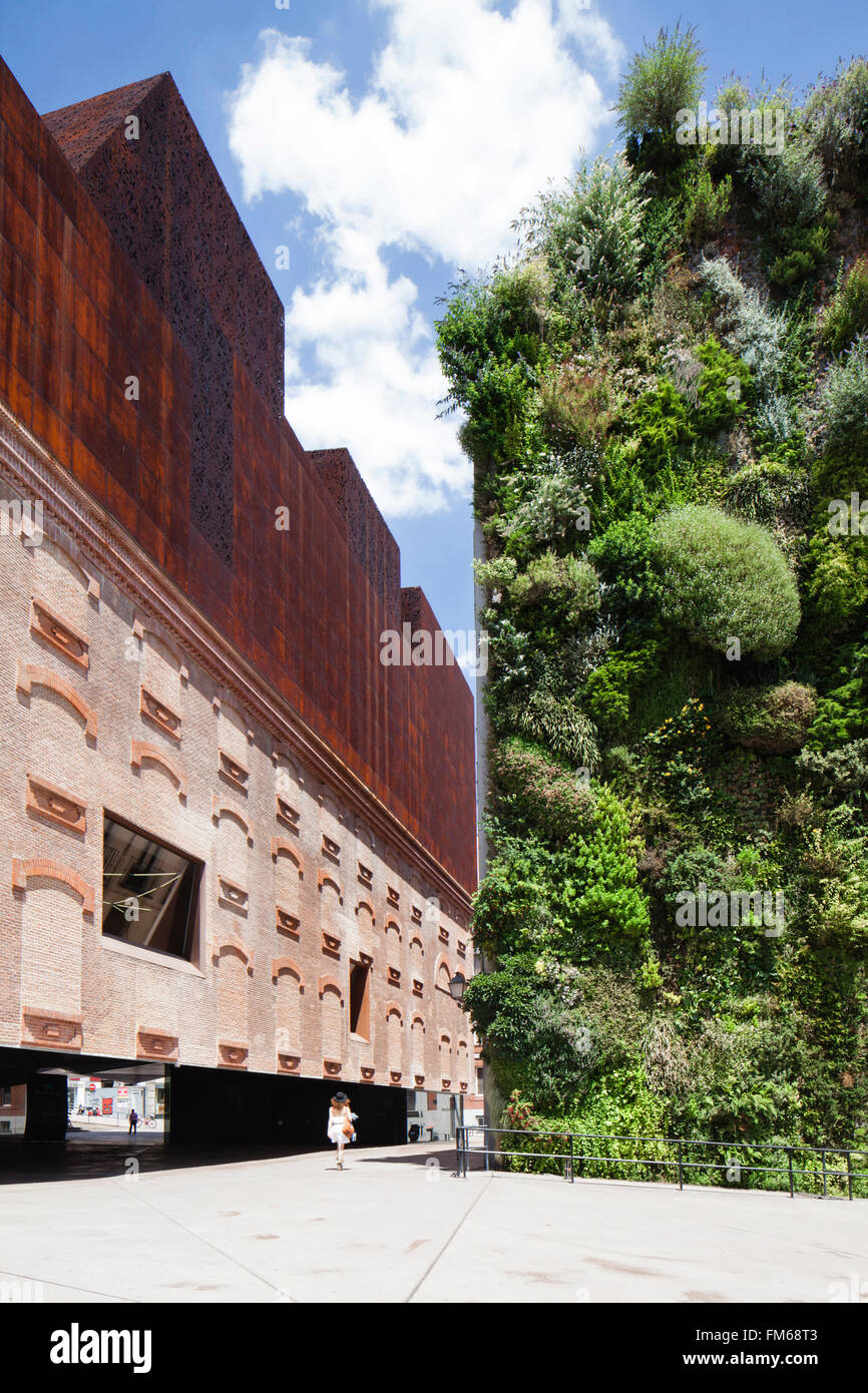 La vue latérale d'un bâtiment appelé La Caixa Forum, à Madrid, avec la conception inhabituelle, comme une dame passe entre elle et un mur de plantes à côté. Banque D'Images