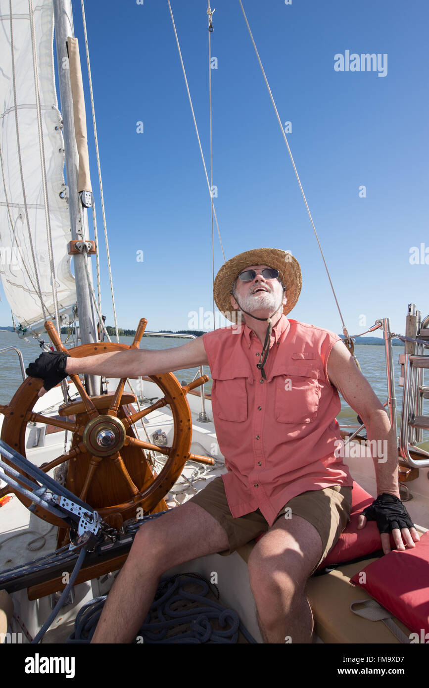 Un homme à la barre de son voilier classique juges le vent dans les voiles sous un ciel bleu. Banque D'Images
