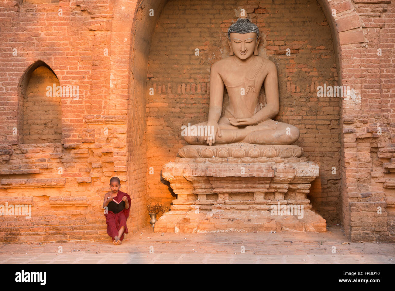 Un jeune moine lecture par une statue de Bouddha, Bagan, Myanmar Banque D'Images