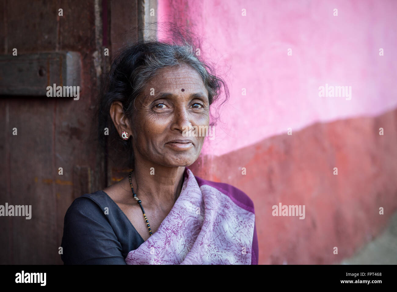 Smiling Indian femme vêtue de pourpre sari rose assis à côté d'un mur à l'extérieur de la maison rurale Banque D'Images
