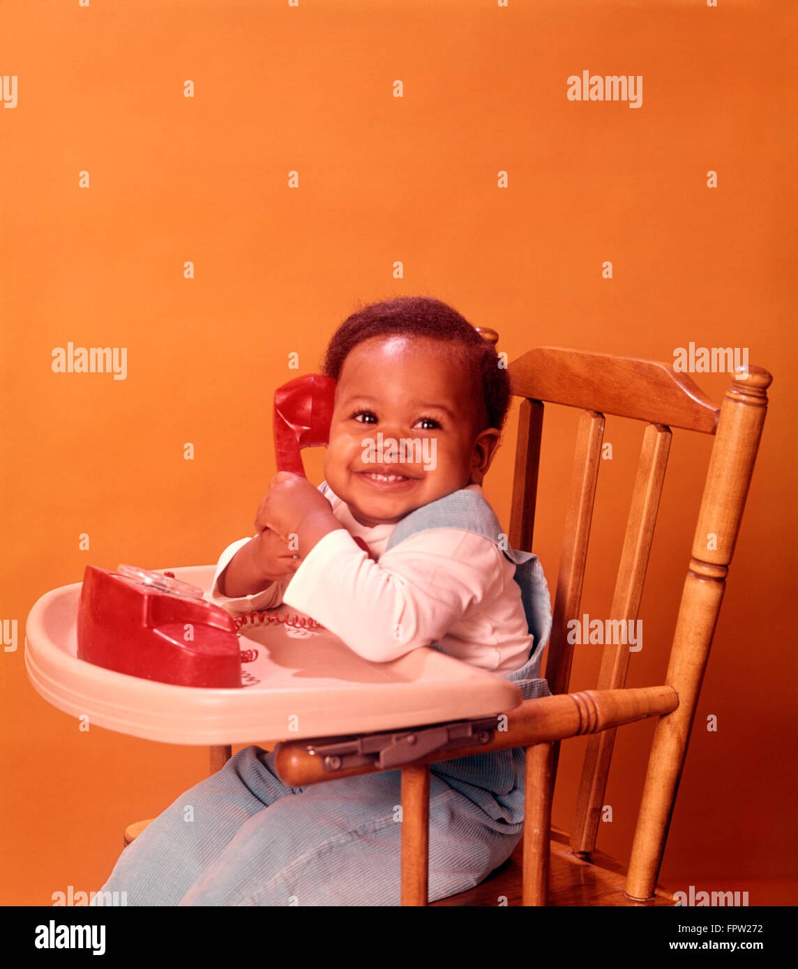 1970 AFRICAN AMERICAN BABY SITTING CHAISE HAUTE BÉBÉ HOLDING TOY ROUGE Téléphone Banque D'Images