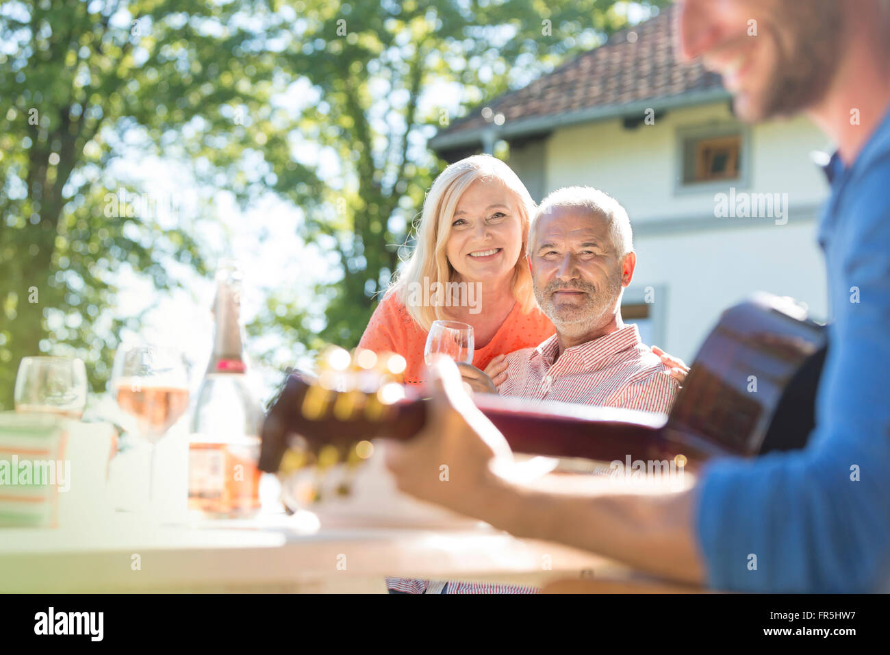 Senior couple drinking wine et regarder des fils adultes à jouer de la guitare sur un patio ensoleillé Banque D'Images