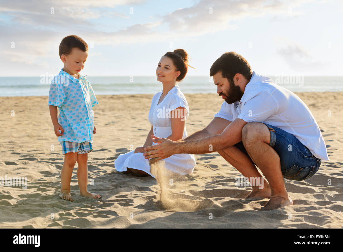Happy Family - mère, père et petit fils bébé assis sur une plage de la mer  au coucher du soleil et jouer avec le sable glisser entre les doigts de la  main