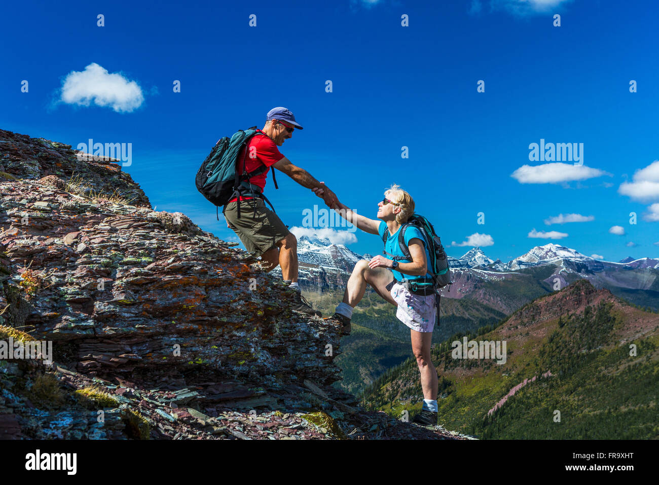 Male hiker aider female hiker jusqu'un pic rocheux d'une crête de montagne avec grand vista et ciel bleu et nuages dans l'arrière-plan Banque D'Images