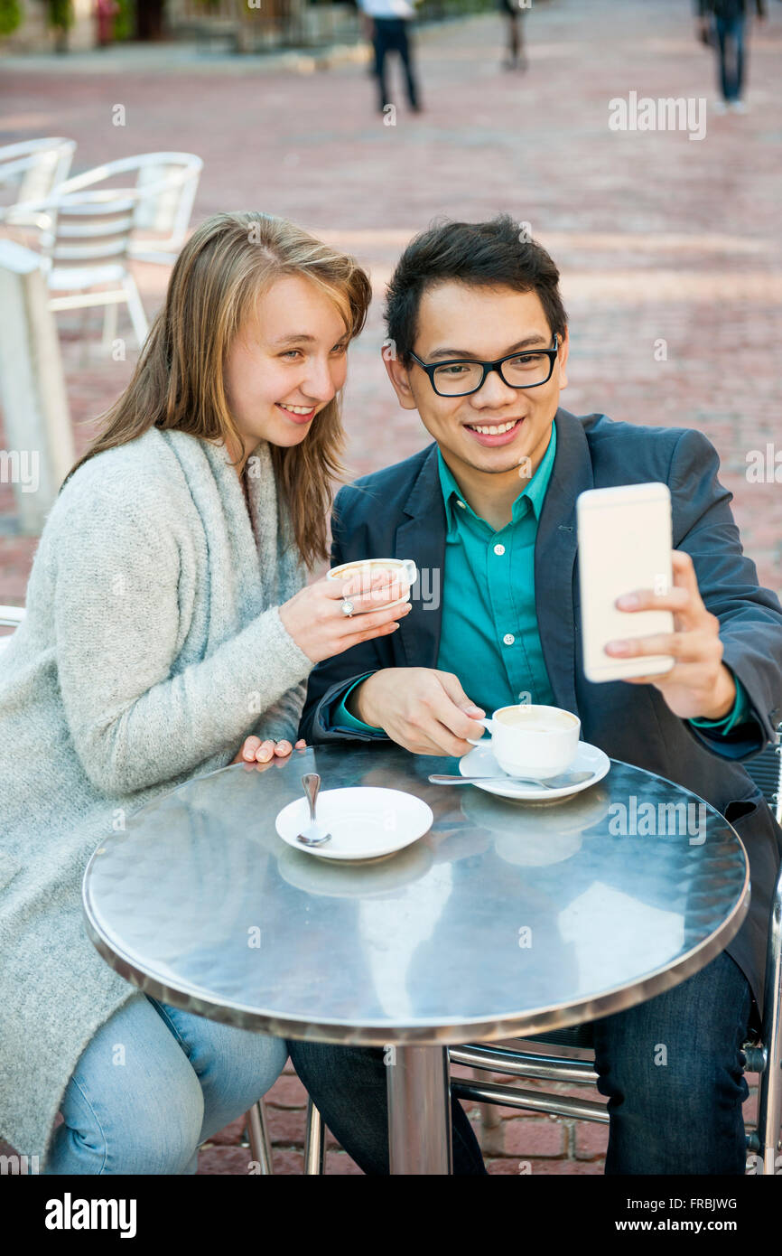 Deux jeunes en souriant à la téléphonie mobile et de prendre une séance tout en selfies at outdoor cafe table Banque D'Images