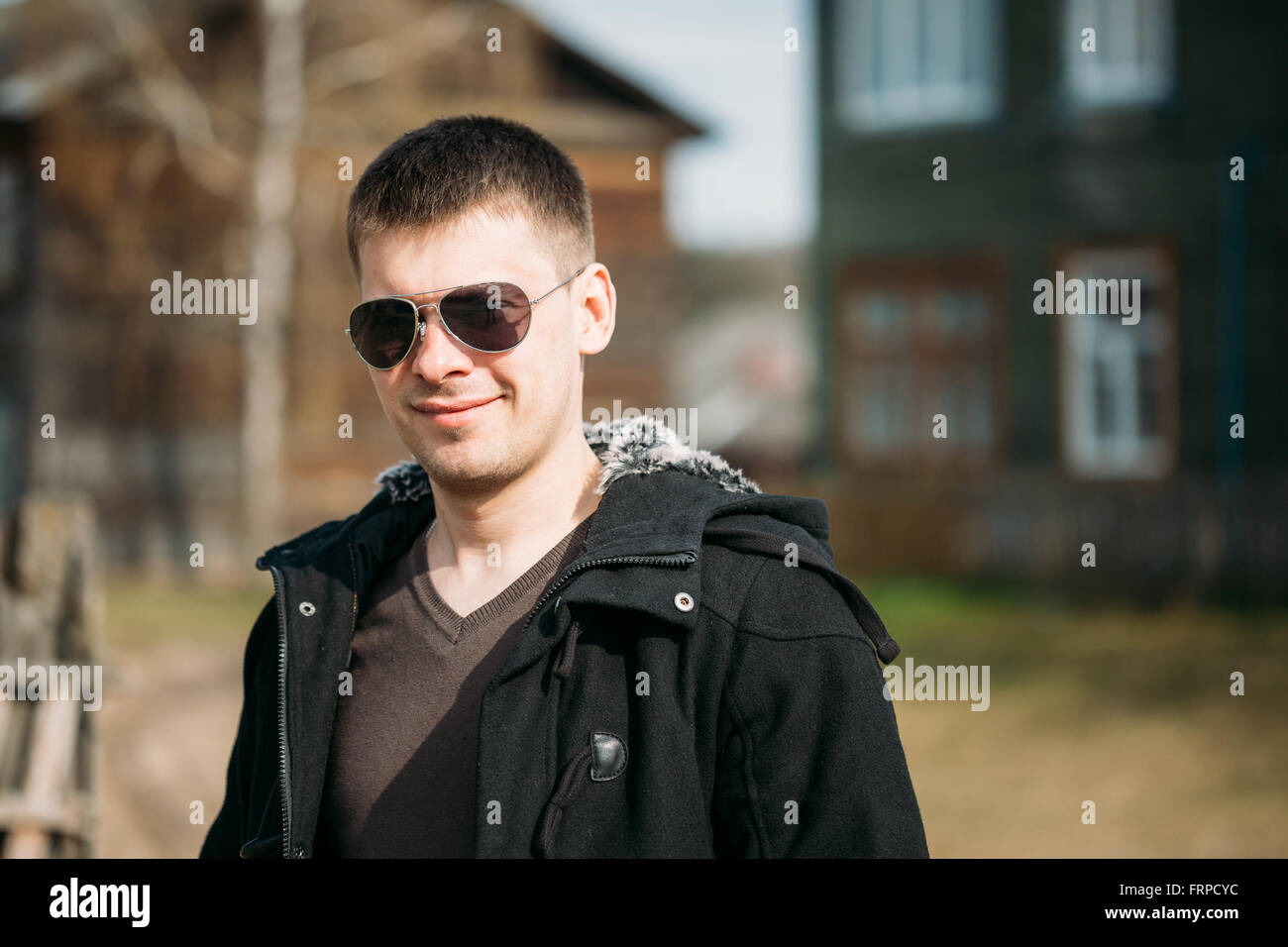 Beau jeune homme d'un séjour près de vieille maison de bois sous le soleil de printemps. Style Casual - veste, lunettes de soleil. Banque D'Images