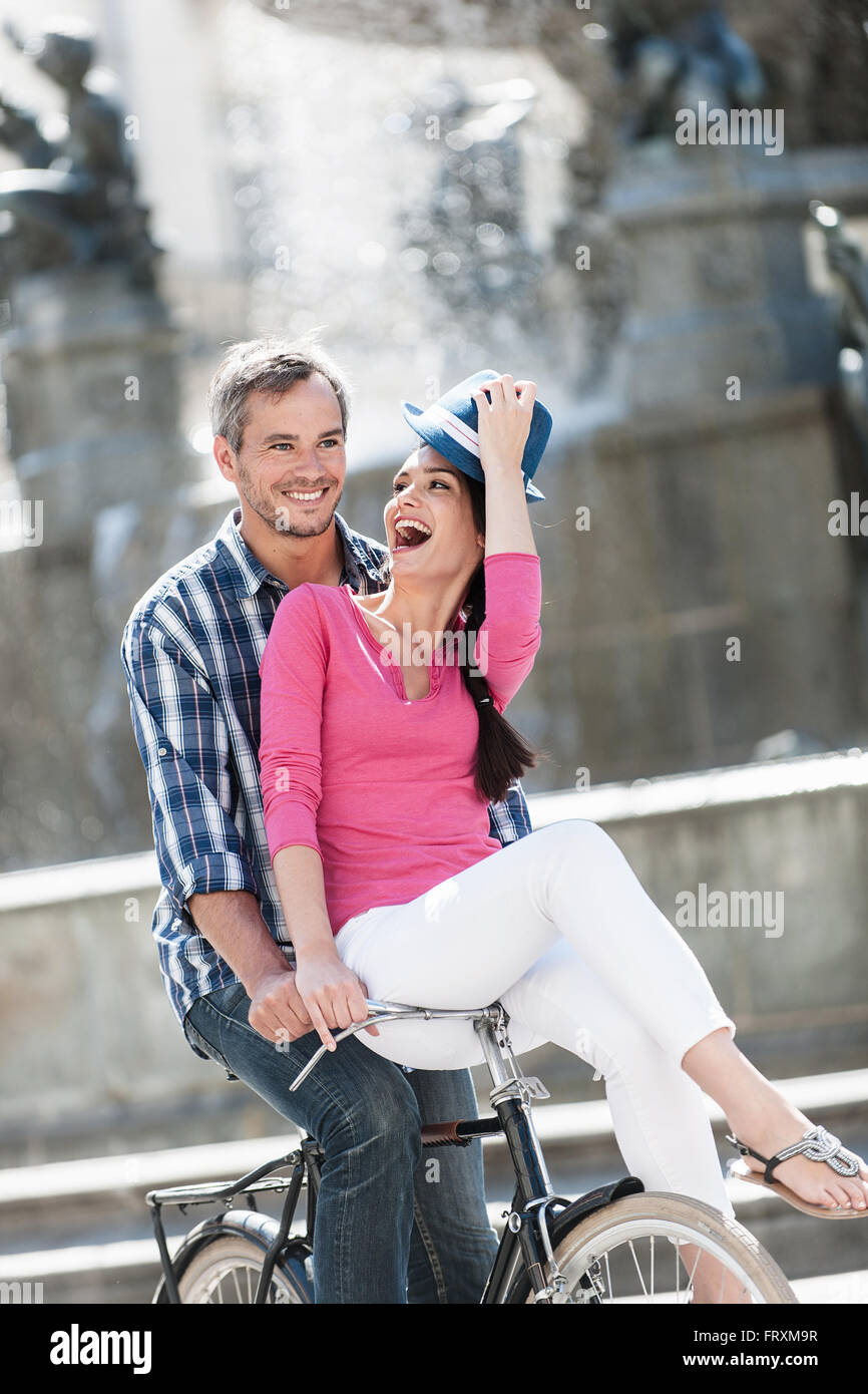 A smiling couple est monté sur un vélo rétro dans le centre-ville. Les cheveux gris l'homme n'est faire du vélo pendant que la femme est assise sur le guidon. Elle porte un haut rose et un chapeau bleu. Banque D'Images
