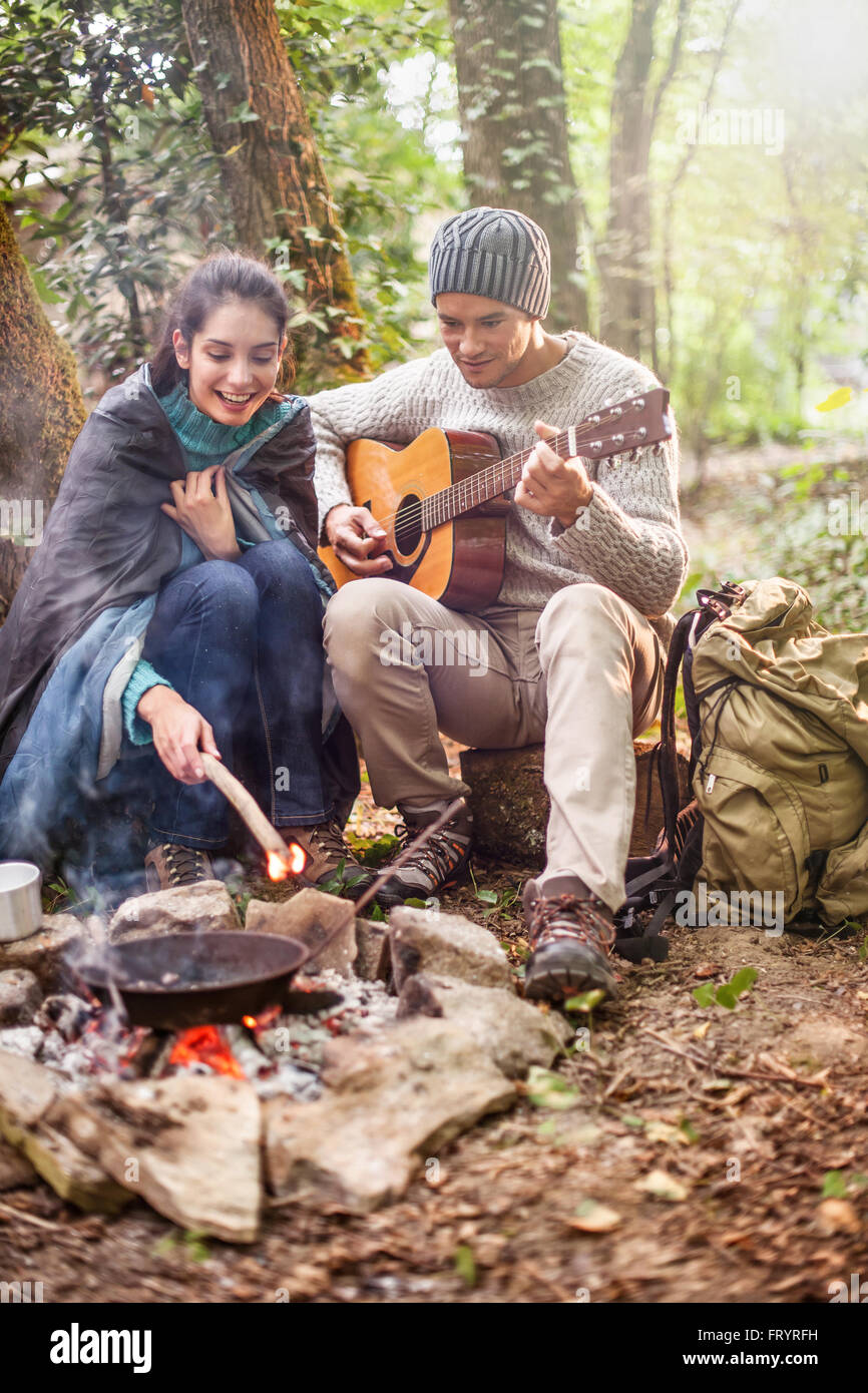 Dans les bois un couple assis près d'un feu de camp, il joue de la guitare,  elle a un sac de couchage sur les épaules pour plus de chaleur Photo Stock -