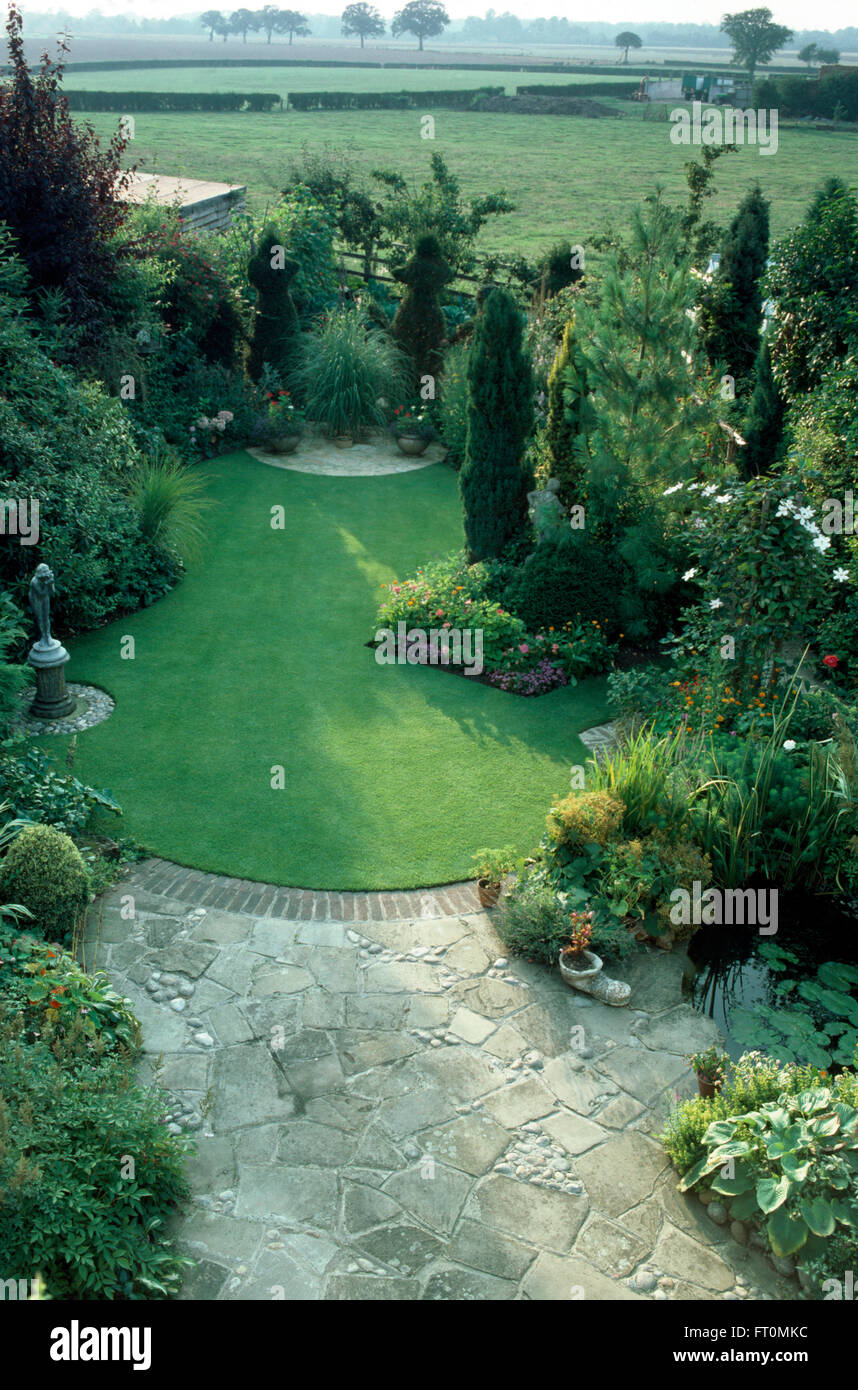 Vue d'un jardin bien entretenu avec un patio pavé fou et une pelouse récemment fauché bordé de conifères et d'arbustes verts Banque D'Images