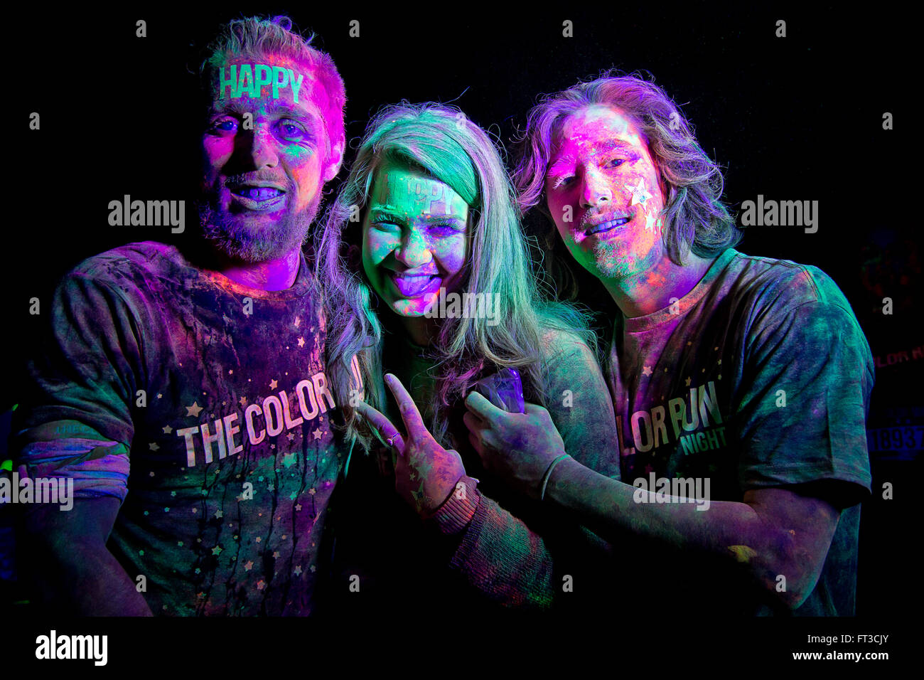Trois adultes prenant part à la Color Run nuit 5K fun run. Elles sont couvertes de poudre de couleur sous la lumière UV. Banque D'Images