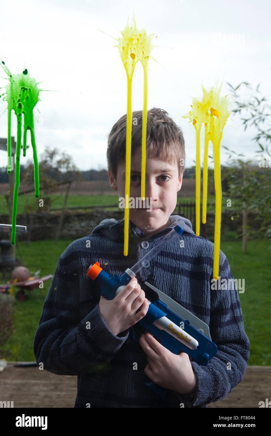 Un jeune garçon se tient fièrement avec son pistolet de paintball, des  éclaboussures de peinture peut être vu sur la fenêtre où il a tiré et c'est  de couler le long de