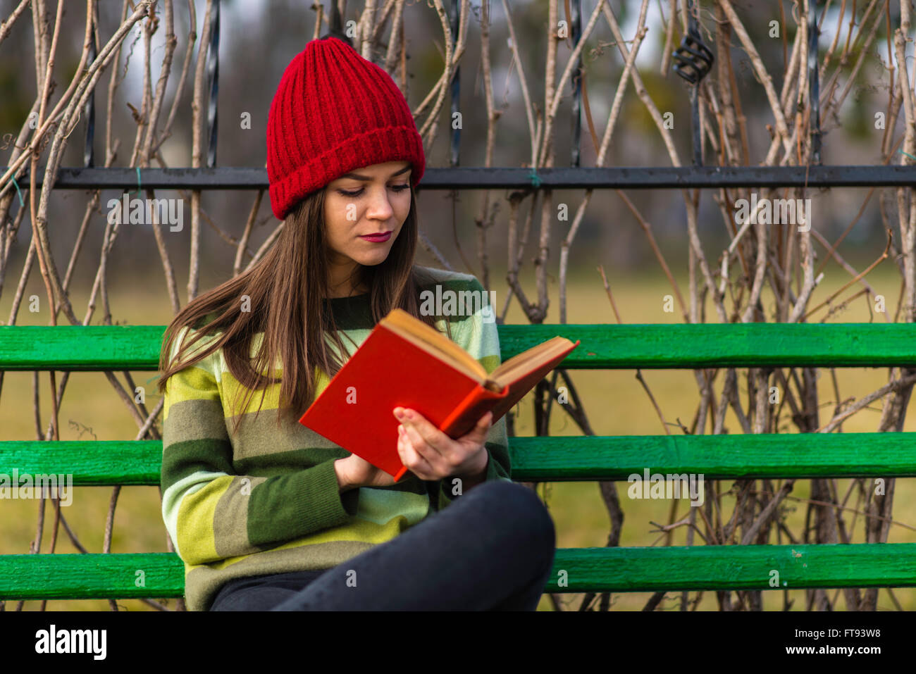 Adolescent Girl in red hat et pull vert assis sur un banc dans un parc et la lecture d'un livre avec couvercle rouge. Banque D'Images